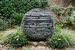 Slate sculpture for garden shown as a hand-stacked slate sphere among boxwood and ornamental grasses